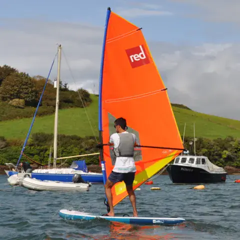 Windsurfeur à la voile orange dans la rade, entre des bateaux au mouillage