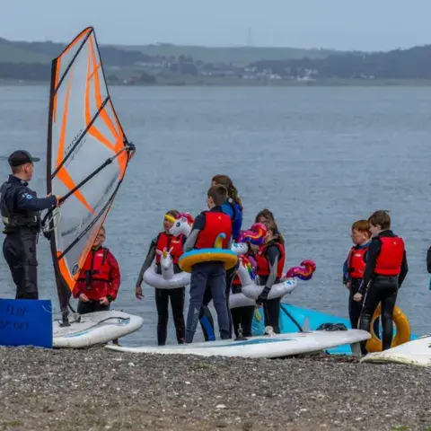Un groupe en gilets sur la plage de galets, avec des planches à voile