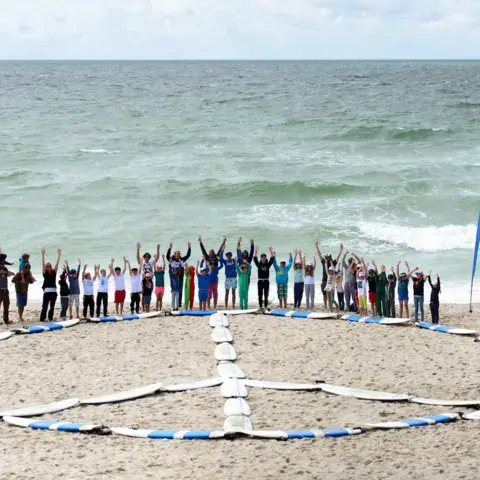 Rassemblement de surfeurs sur la plage, signe de paix dessiné avec des planches