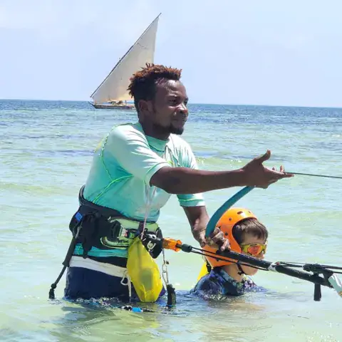 Moniteur guidant un élève dans l'eau, avec un boutre au loin