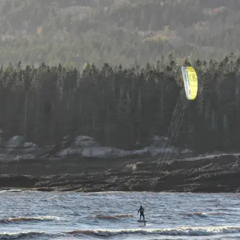 Un kitesurfeur sous une aile jaune, au pied d'une côte boisée