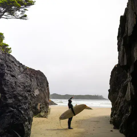 Surfeur avec sa planche entre deux rochers sur une plage de Tofino