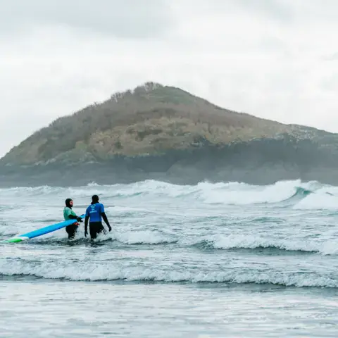 Deux surfeurs entrant dans l'eau devant une île rocheuse