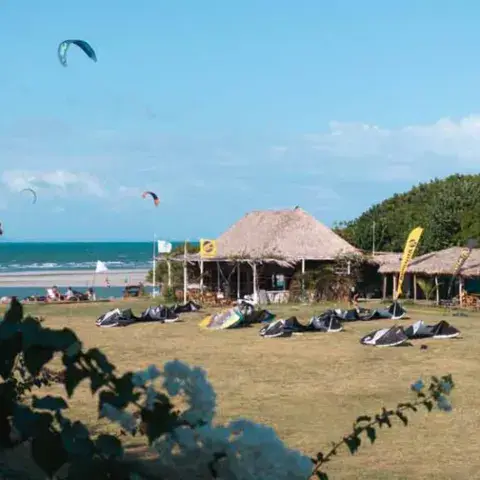 Terrain herbeux avec des kites posés au sol et des paillotes, plage derrière