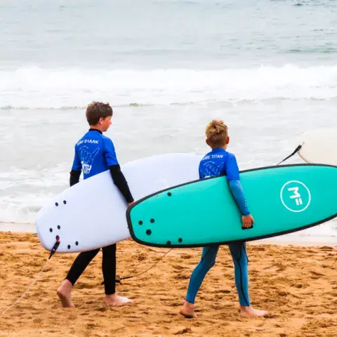 Deux élèves de dos portant leurs planches sur la plage