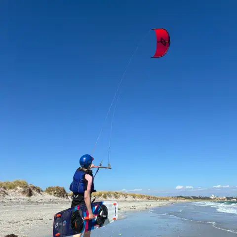 Élève avec sa planche sur la plage, kite dans le ciel
