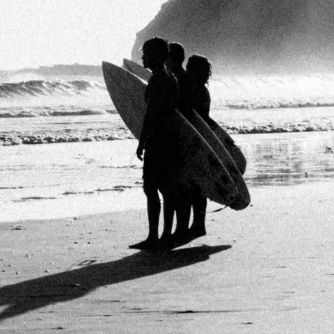 Trois surfeurs avec leurs planches au bord de l'eau, en noir et blanc