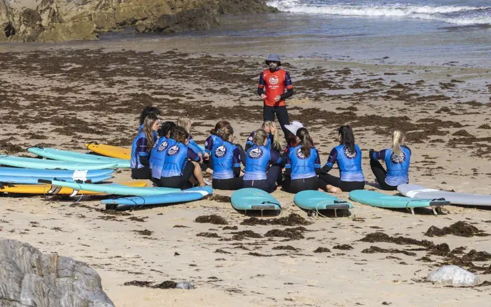 Groupe d'élèves assis avec leurs planches sur la plage, face au moniteur