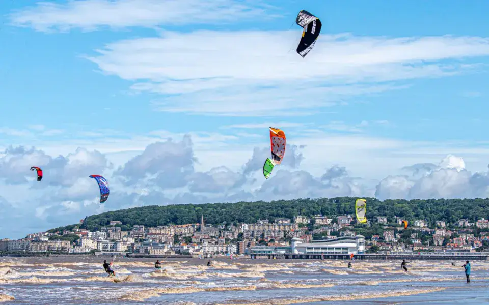 Nombreux kitesurfeurs au-dessus d'une plage, devant une ville