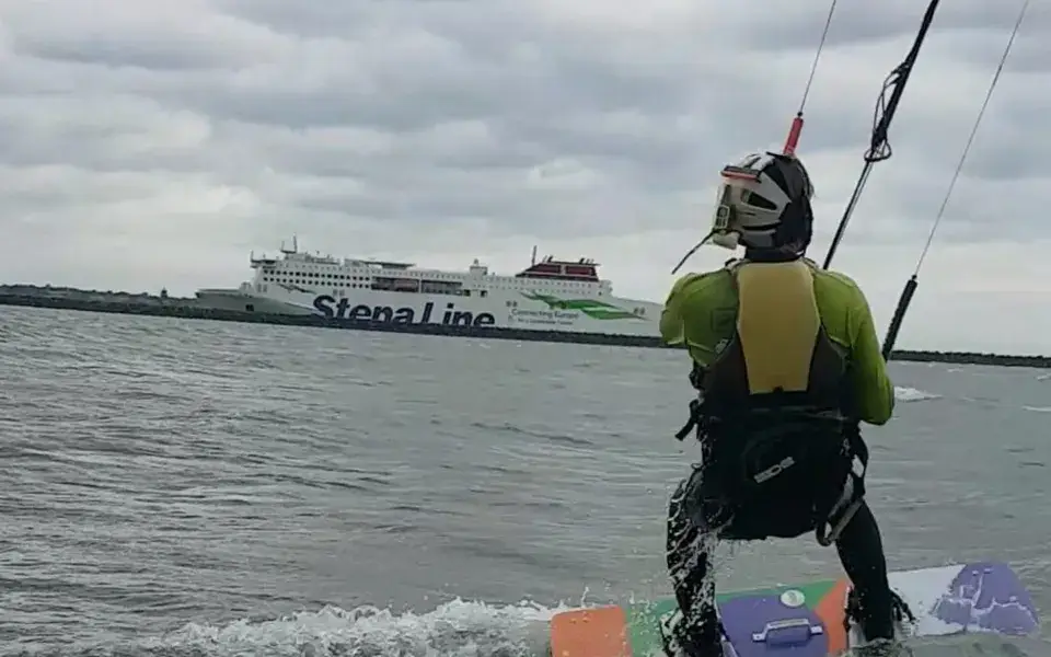 Élève de kitesurf sur sa planche, vu de dos, près d'un ferry