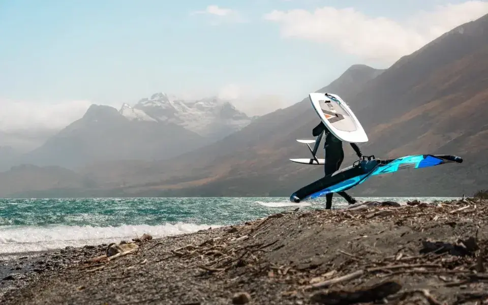Wingfoileur portant sa planche et son aile sur une rive de galets, montagnes derrière