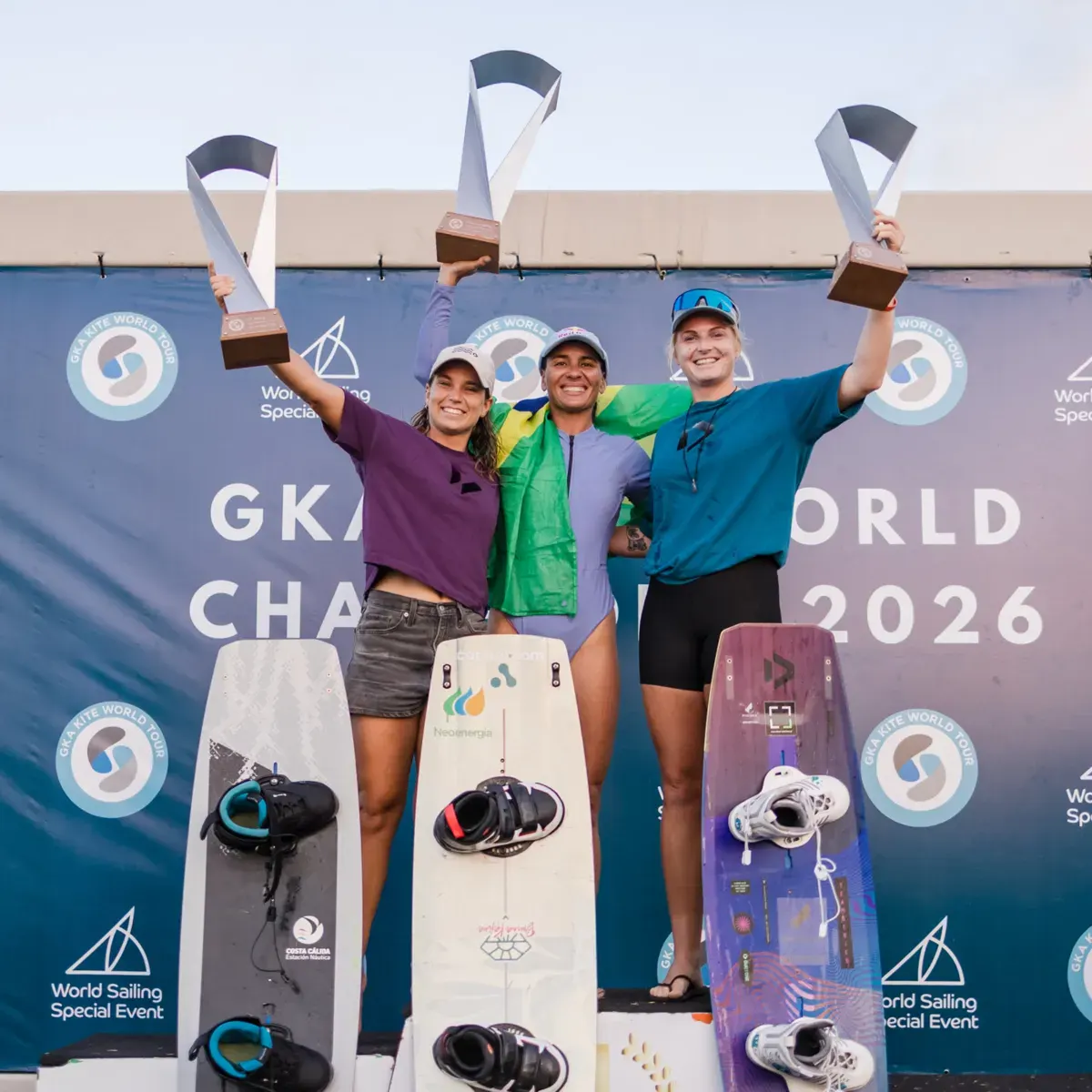 Le podium féminin d'Ilha do Guajiru, trophées levés, avec Bruna Kajiya au centre drapée dans le drapeau brésilien