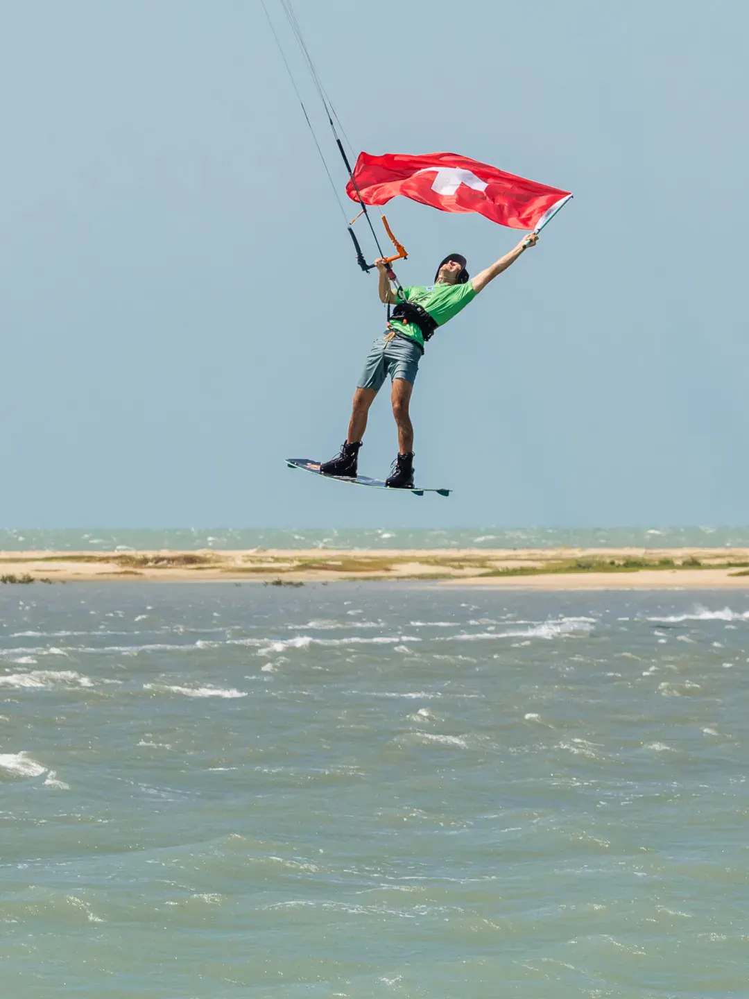 Un rider en plein saut brandit un drapeau suisse au-dessus du lagon d'Ilha do Guajiru