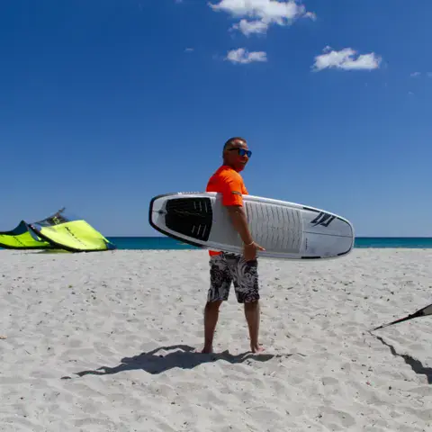 Moniteur portant une planche de kitesurf sur la plage, kites au sol en arrière-plan