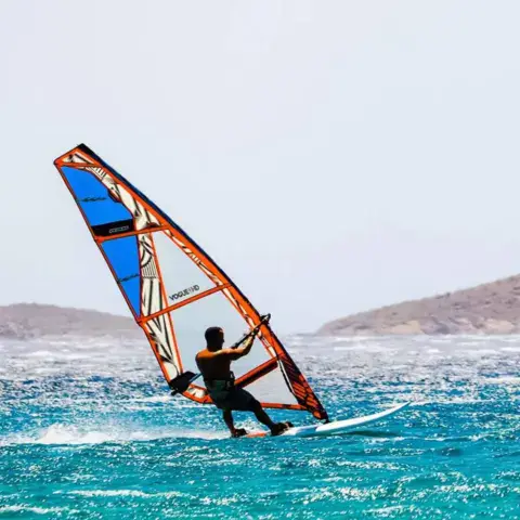 Windsurfeur sous une voile orange et bleue devant la côte
