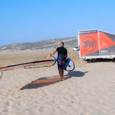 Windsurfeur portant son gréement sur la plage, remorque de la station au fond