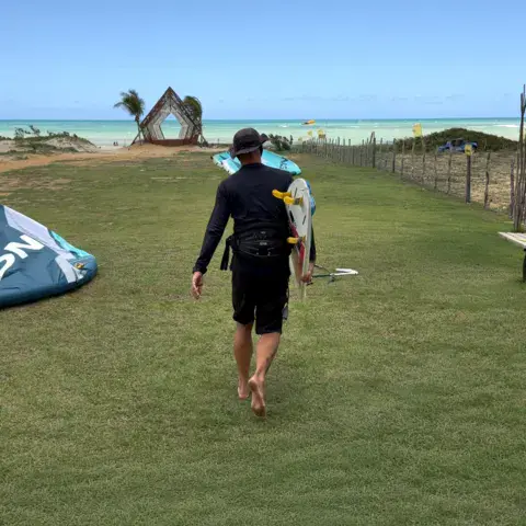 Kiter portant son kite et sa planche sur l'herbe, plage en arrière-plan