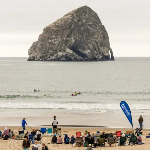 Plage avec un gros rocher au large et un rassemblement de surfeurs