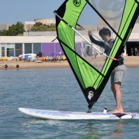 Élève debout sur une planche à voile, base nautique en arrière-plan