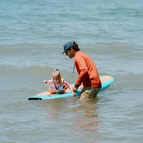 Moniteur poussant un jeune enfant allongé sur une planche, dans de petites vagues