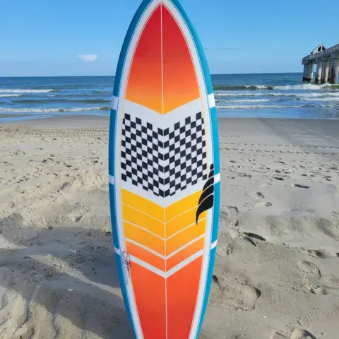 Planche de surf orange et bleue plantée dans le sable, avec la jetée au fond