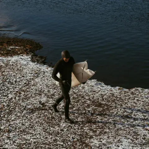 Surfeur en combinaison portant sa planche sur une rive enneigée du lac