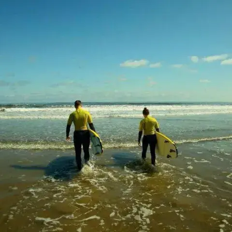 Deux élèves en haut jaune entrant dans l'eau avec leurs planches