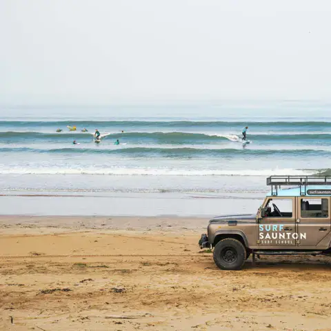 Land Rover de l'école sur la plage, avec des surfeurs dans les vagues