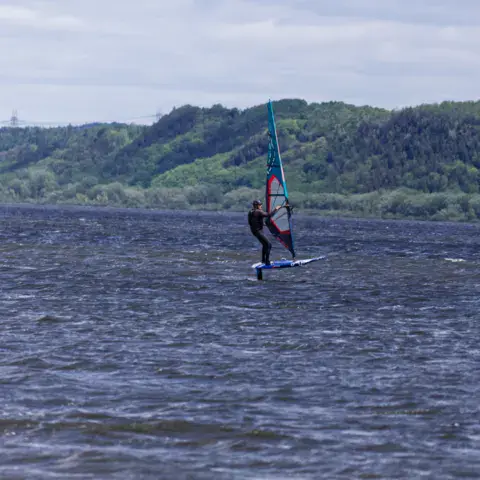 Planche à voile sur l'eau agitée, collines boisées au fond