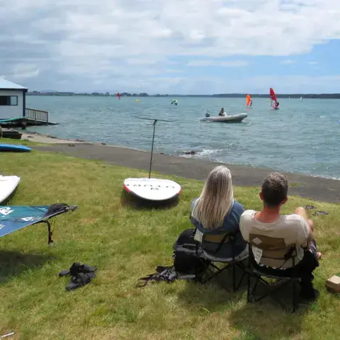 Le hangar du club et des planches sur l'herbe, face au plan d'eau