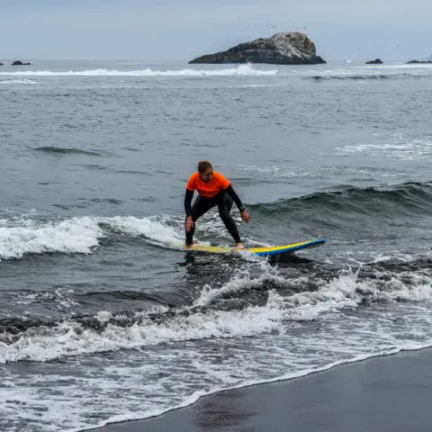 Surfeur en combinaison orange sur une vague, rocher au fond